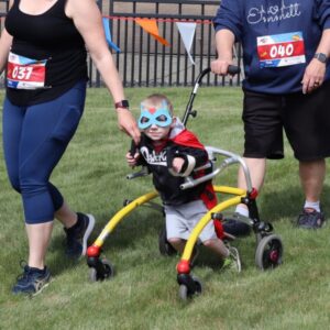 A young boy wearing a light blue superhero eye mask, walks on grass with the use of a mobility walker. On either side of him are his parents.