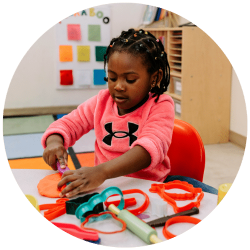 Child sitting at a table in their classroom plays with a cookie cutter and playdoh