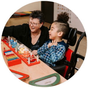 Child in a wheelchair playing a xylophone with his classroom aide.