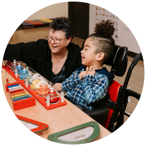 Child in a wheelchair playing a xylophone with his classroom aide.