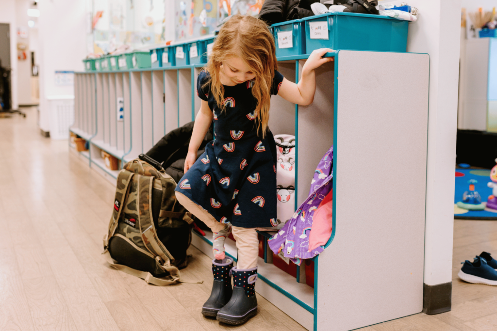 A young girl puts on boots with a wall of open lockers behind her