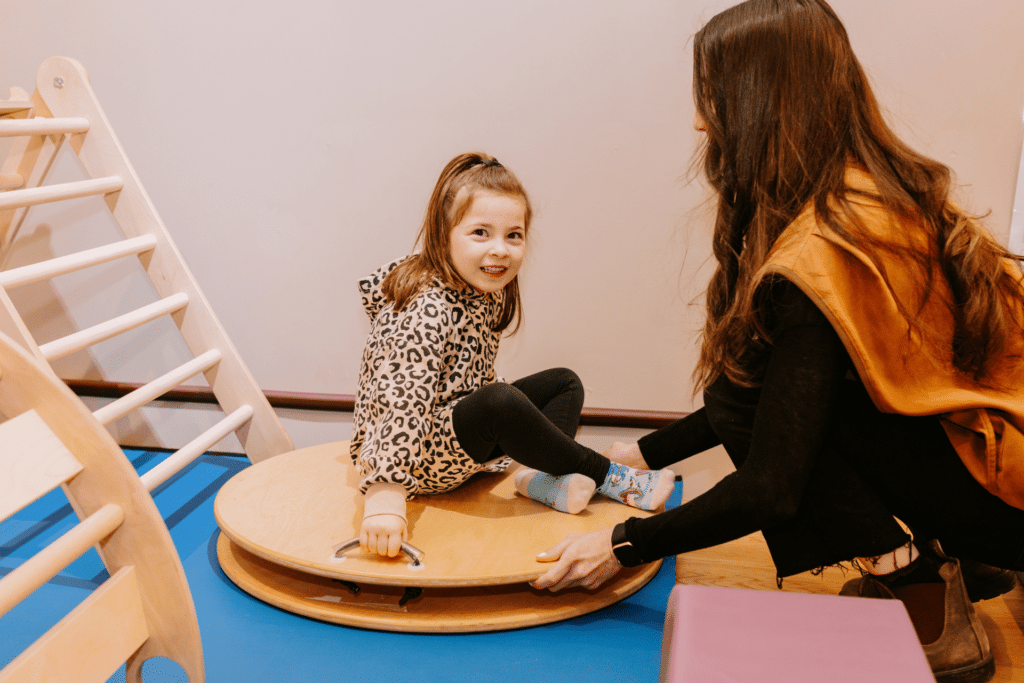 A young girl is spun by her class aide on a round, wooden wheel