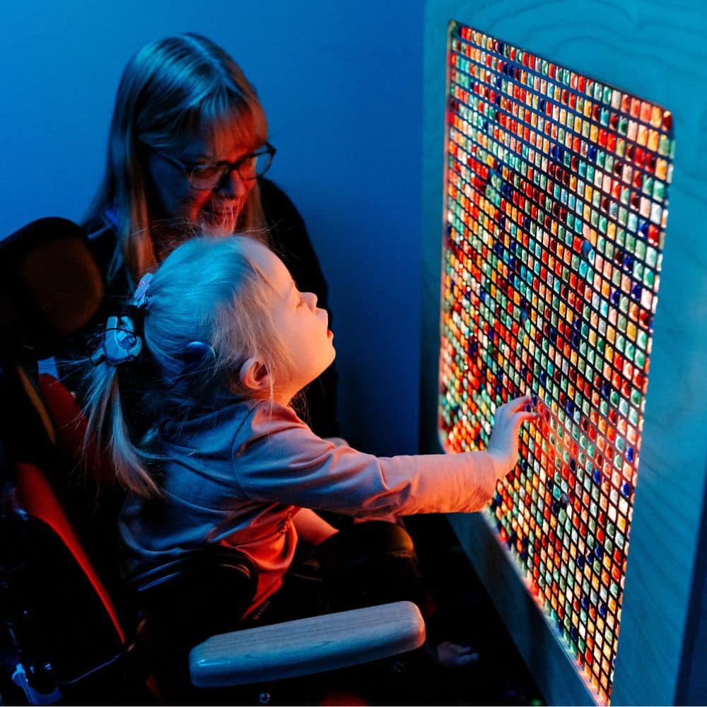 A young girl in a wheelchair touches a light wall in a dark sensory room. An aide worker kneels beside her.
