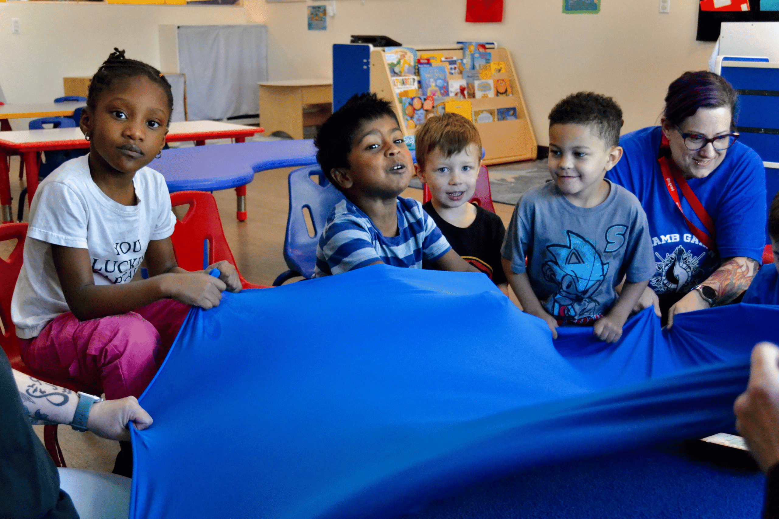 A group of ethically diverse young boys with diverse needs and a classroom therapy aide with their hands up in a classroom setting play the parachute game